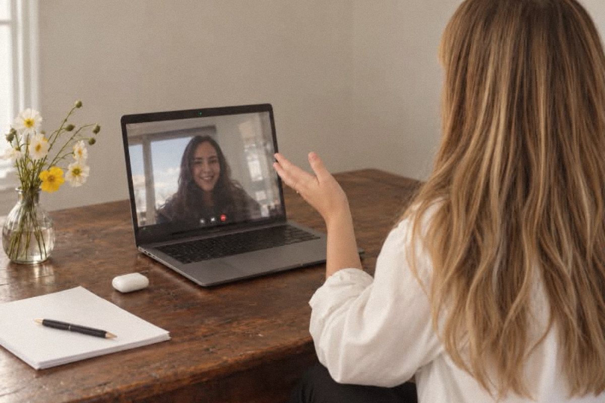 A person at a wooden desk in an NDIS telehealth psychology session, waving to a smiling psychologist on screen
